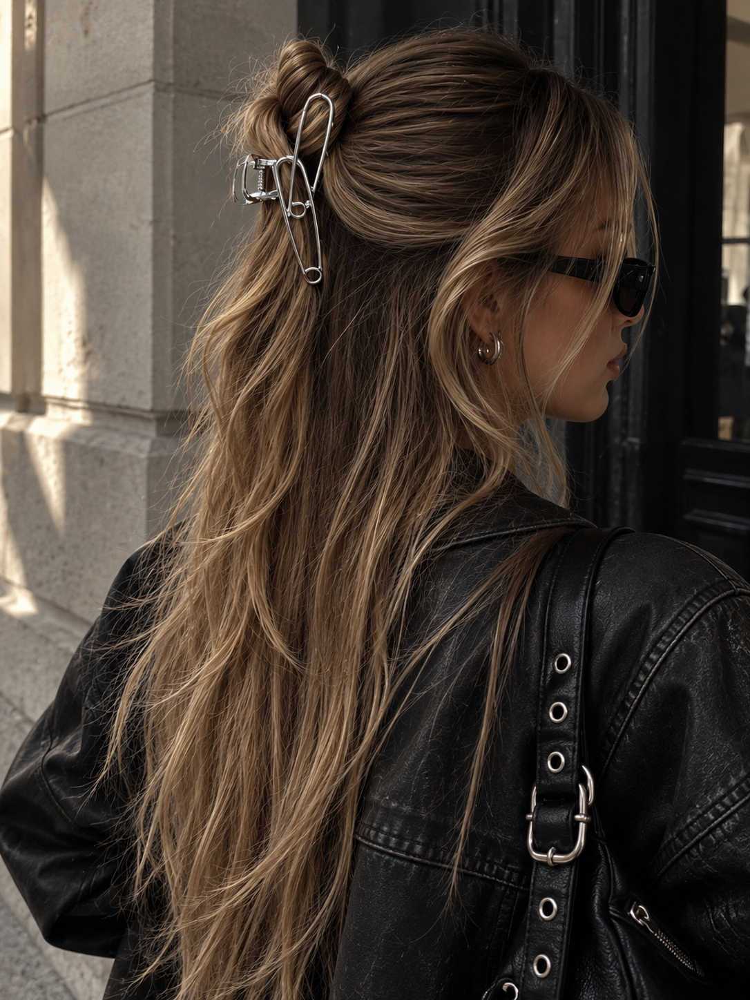 A woman shown from the back wearing a large silver safety-pin style hair claw clip holding a half-up ponytail. She has long highlighted brown hair, wears a black leather jacket, and is photographed outdoors against a stone building and dark doorway.